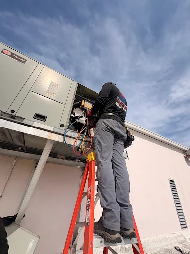 Technician on a ladder servicing a rooftop packaged AC unit against a blue sky