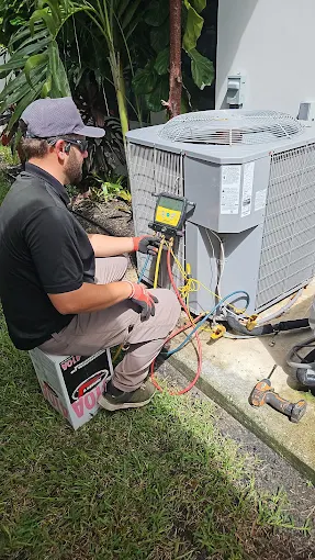 Technician servicing an outdoor AC condenser with a digital manifold gauge beside tropical landscaping