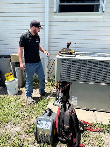Technician servicing an outdoor AC condenser with a recovery machine and manifold gauges on a lawn