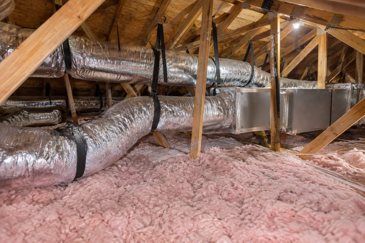 Attic of a Florida home with new silver insulated flex ducts neatly strapped and a sealed sheet-metal trunk line, over pink insulation, lit by work lights