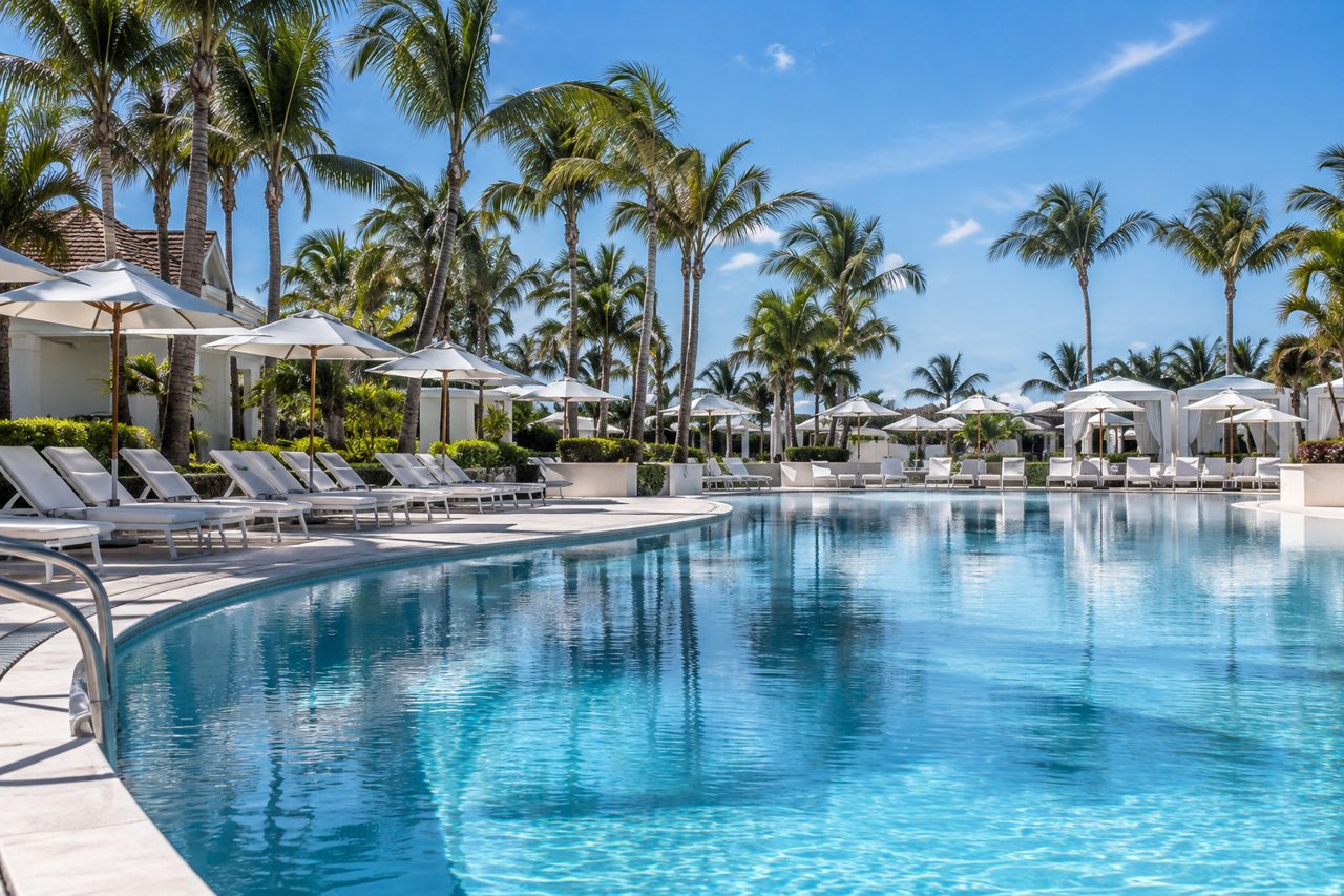 Large resort pool in Palm Beach Gardens with rows of white loungers and umbrellas, palms, blue sky, no people